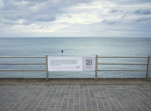 A board attached to the beach wall at Margate, with the statue visible in the distance beyond it. The board contains a detailed image description of the statue and the RNIB logo